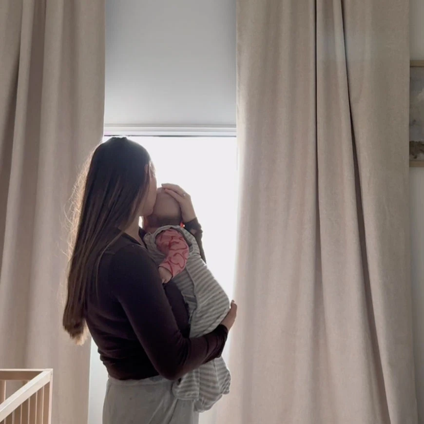 A parent settling a baby by a window with the shade lowered
