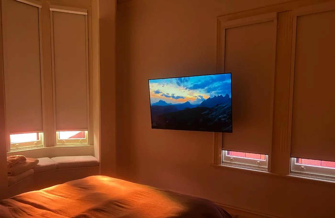 A bedroom at dusk with the shades down and the TV the only light in the room